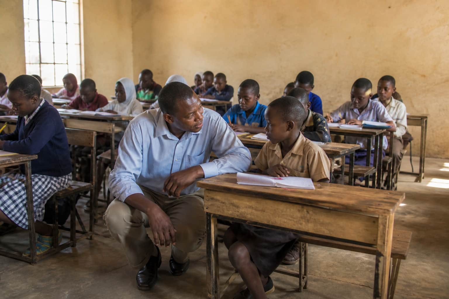 A teacher kneeling beside a student's desk, listening carefully