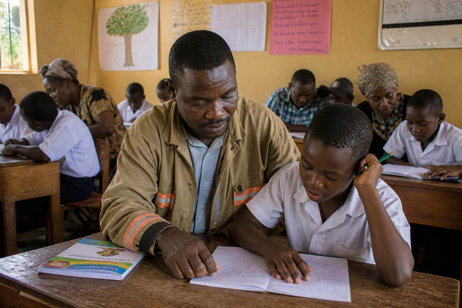 A parent sitting next to their child at a school desk, looking at their work together