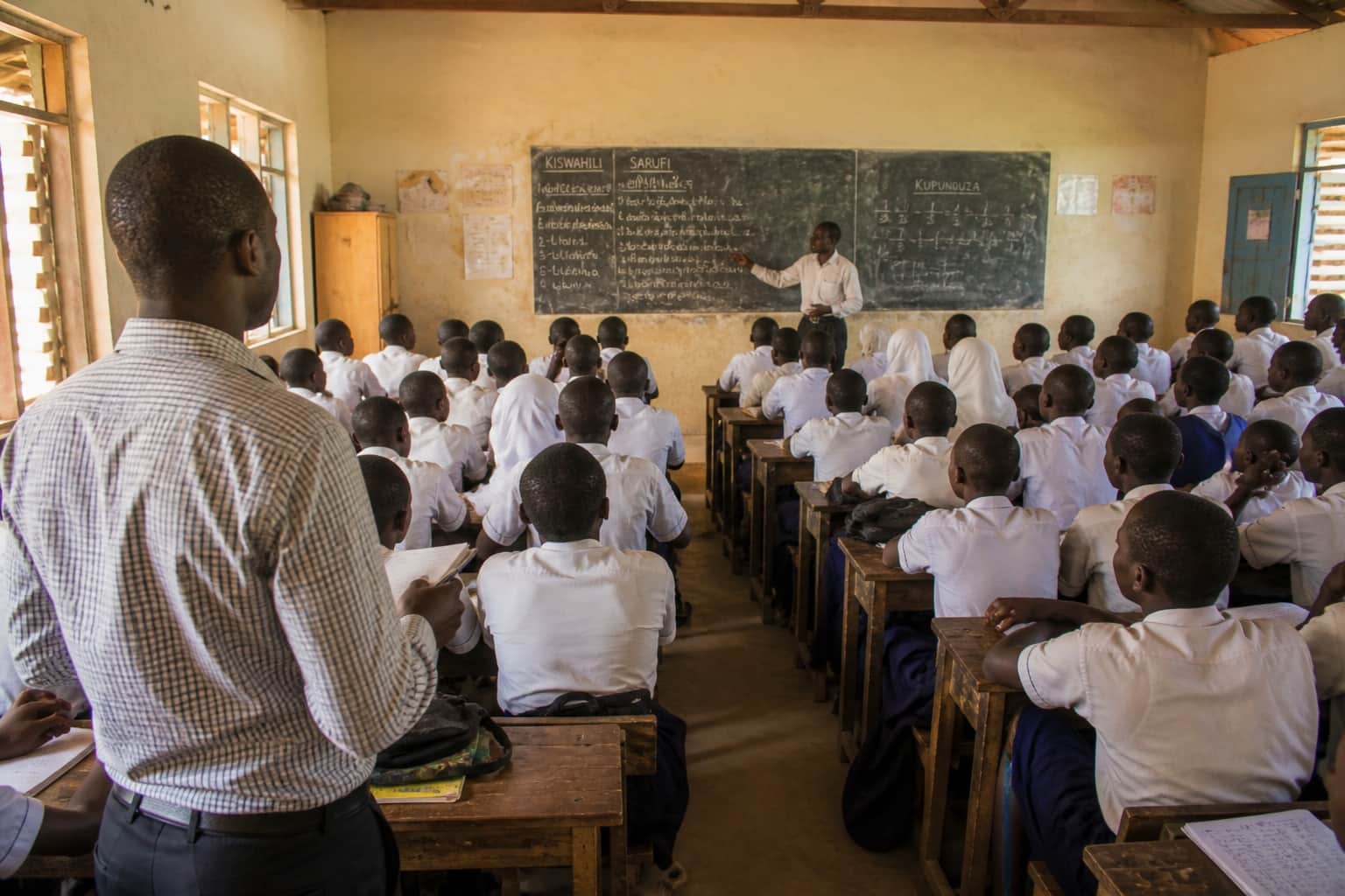 A teacher stands at the back of a busy classroom, watching a colleague teach
